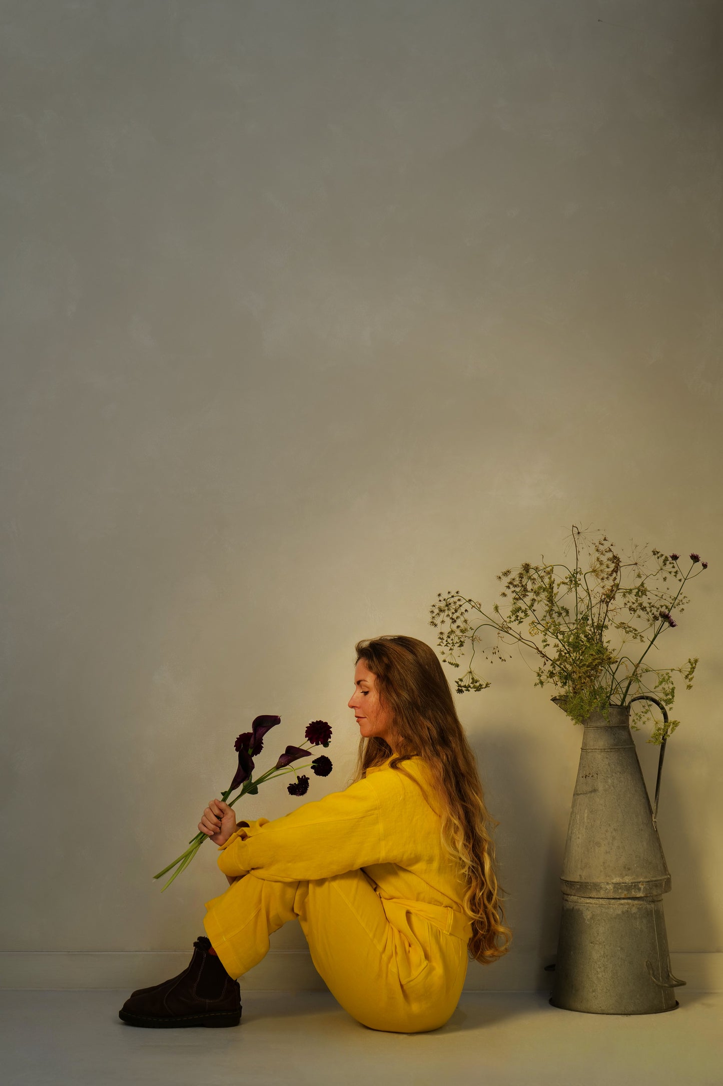 Woman in a yellow linen outfit sitting on the floor with flowers against a lime washed wall as the warm sun set beams in