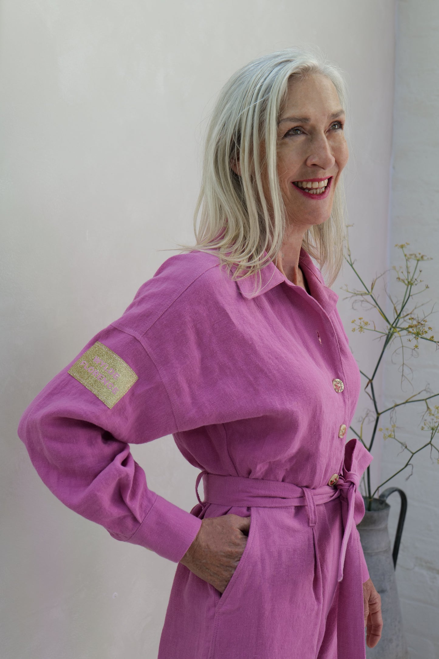 Woman wearing a pink dress with a gold 'wylde florence' patch, standing against a lime washed background.