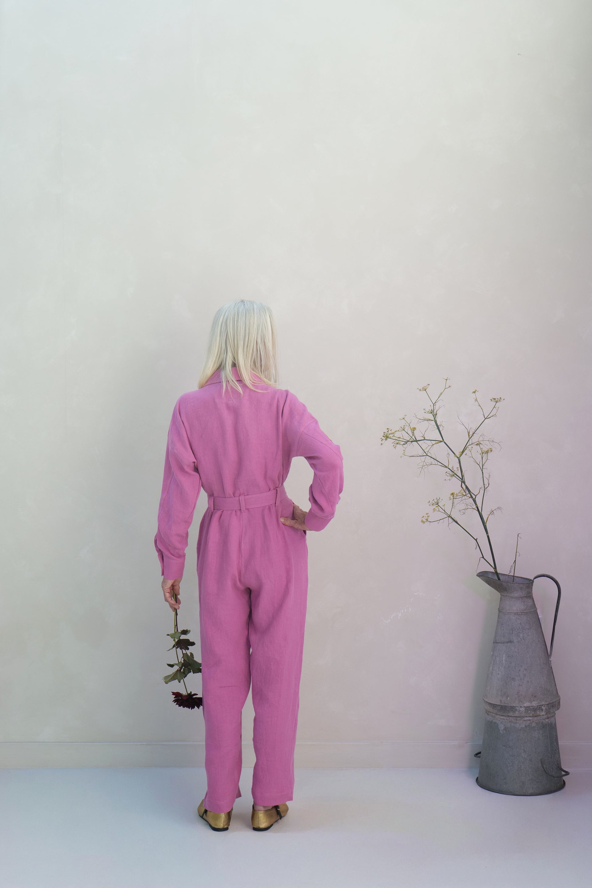 Person wearing a pink linen jumpsuit standing in front of a white wall with a metal pitcher and branches.