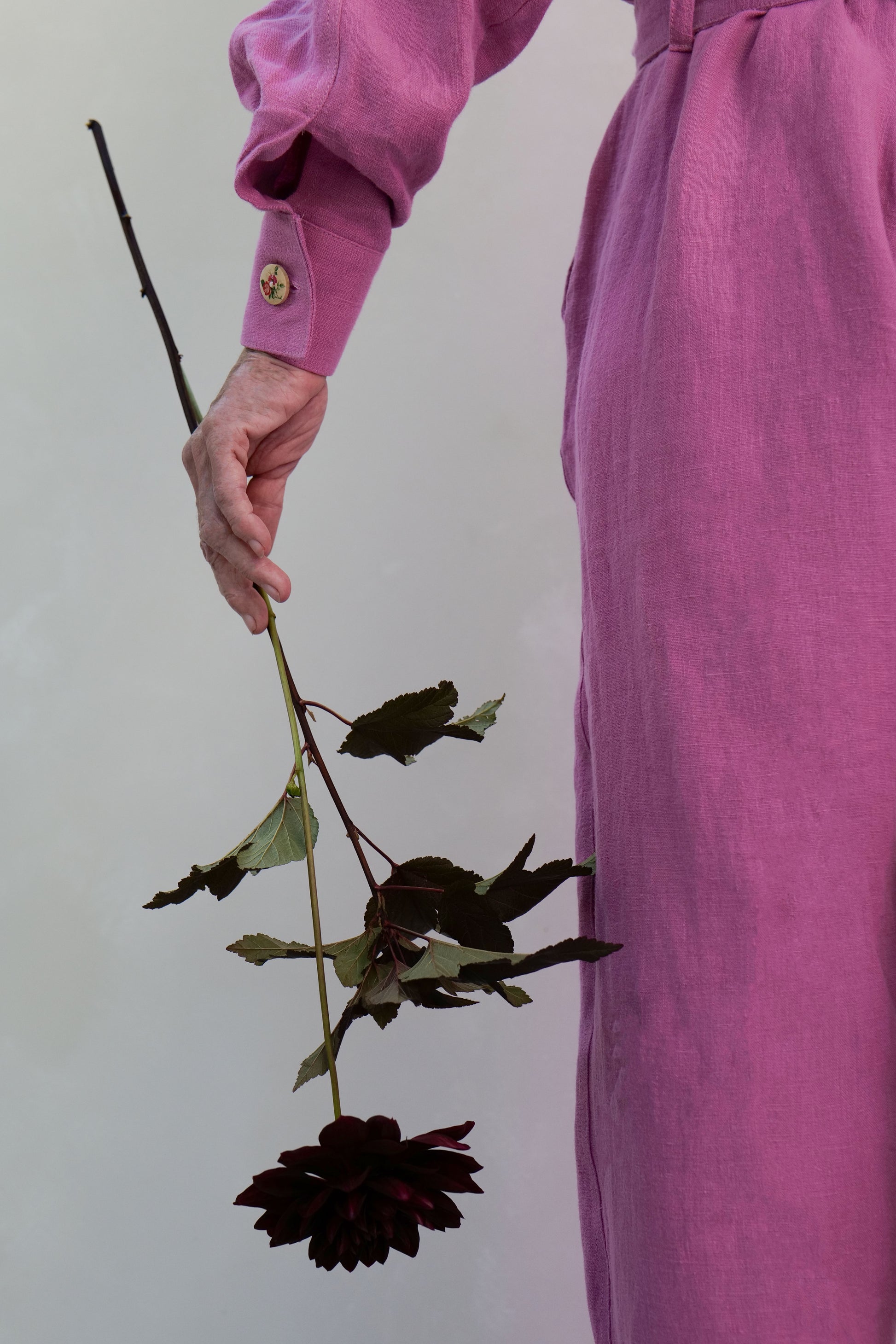 Person in pink linen jumpsuit with floral painted button at cuff, holding a deep red dahlia stem