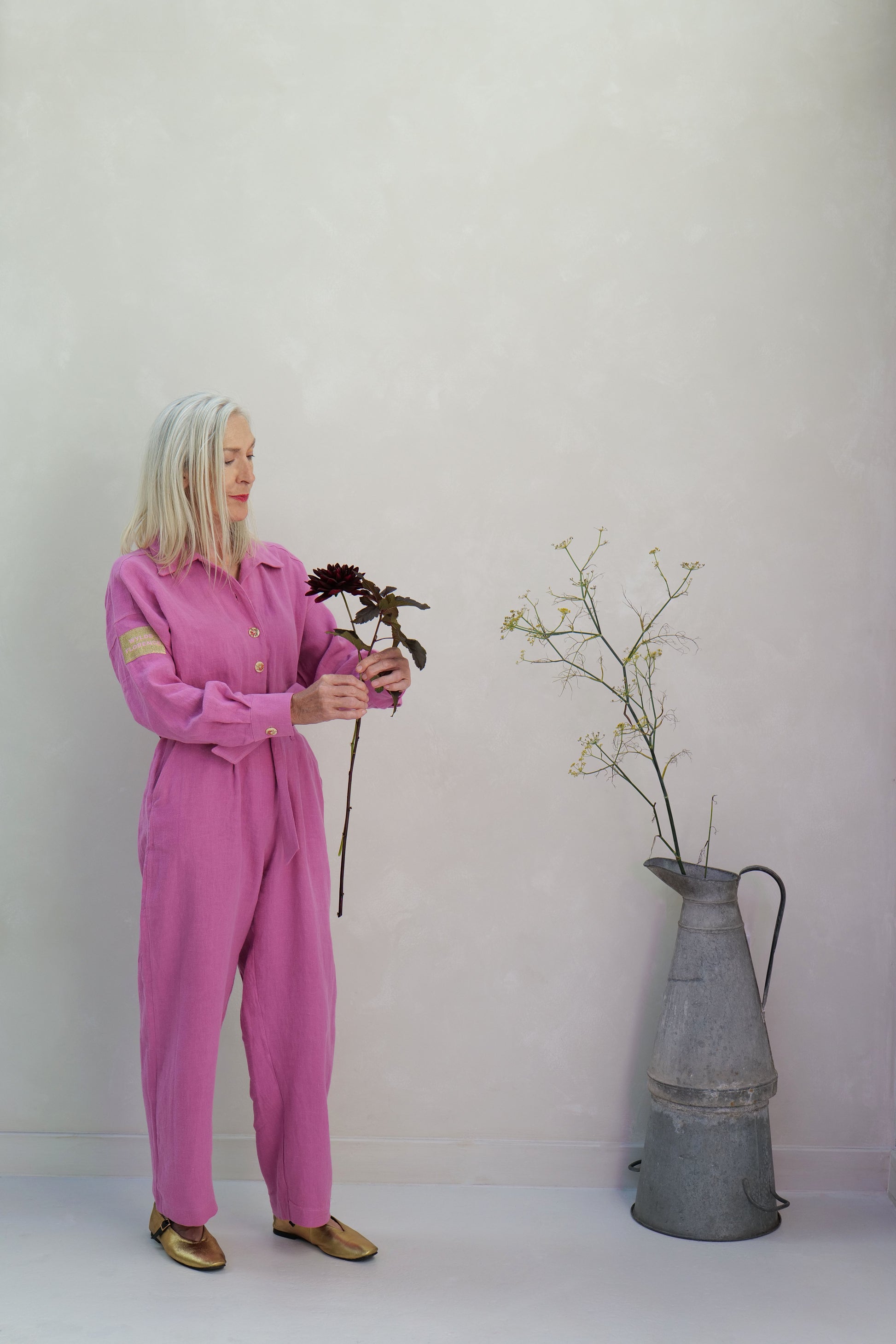 Person in a pink jumpsuit holding flowers next to a metal vase against a white background