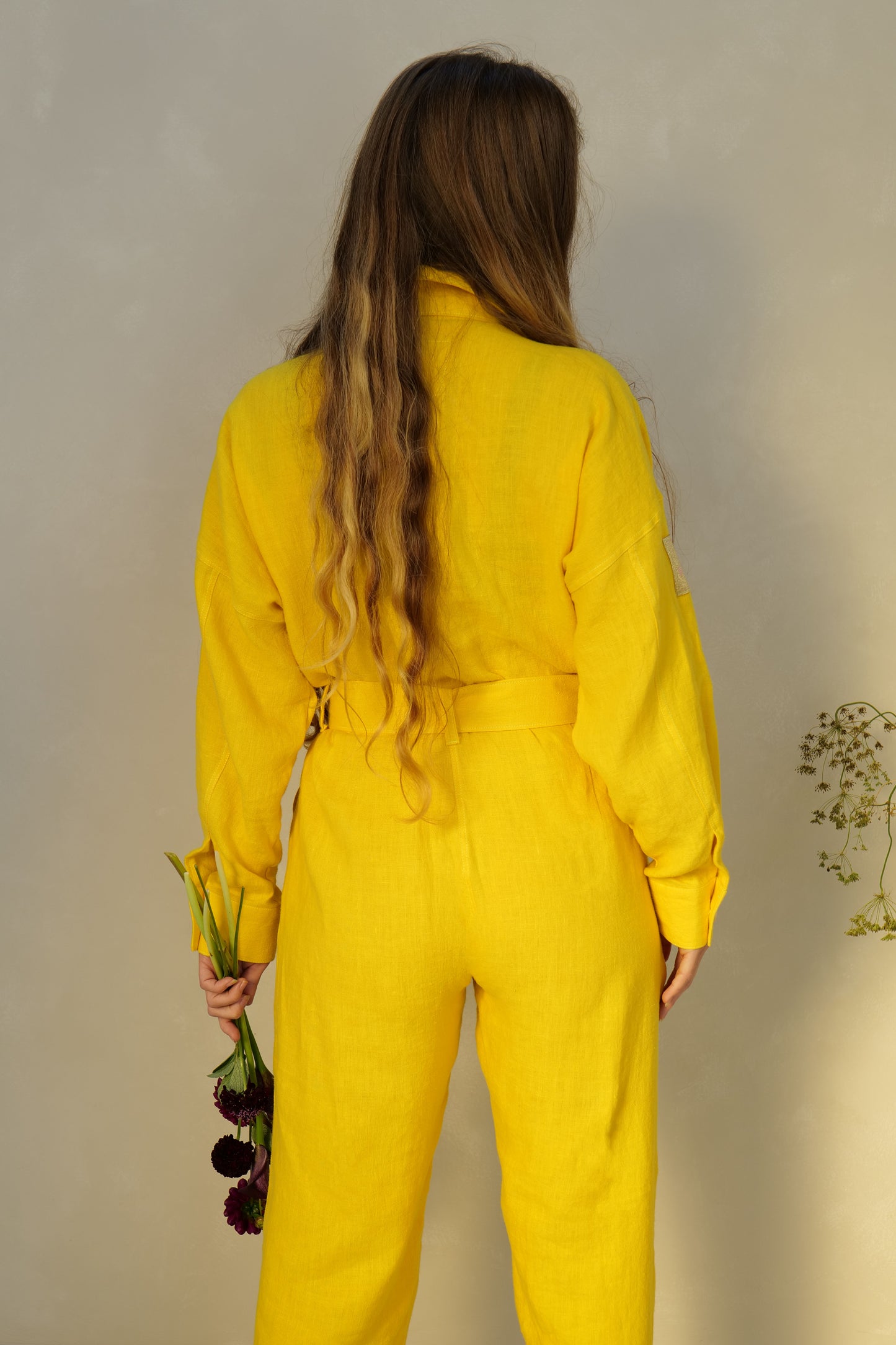 Person wearing a yellow linen jumpsuit holding rich dark red, almost black flowers against a lime washed  background
