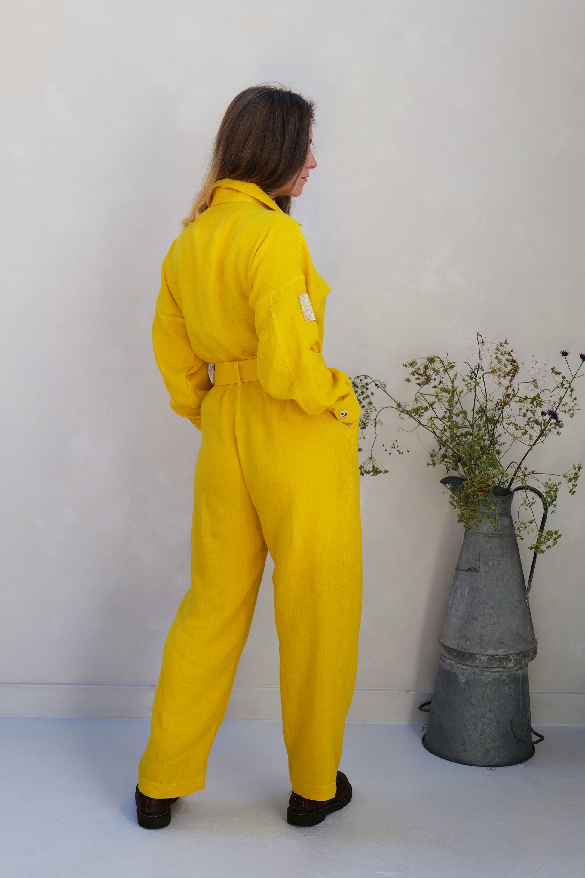 Person wearing yellow linen overalls standing next to a metal vase with plants against a white wall.