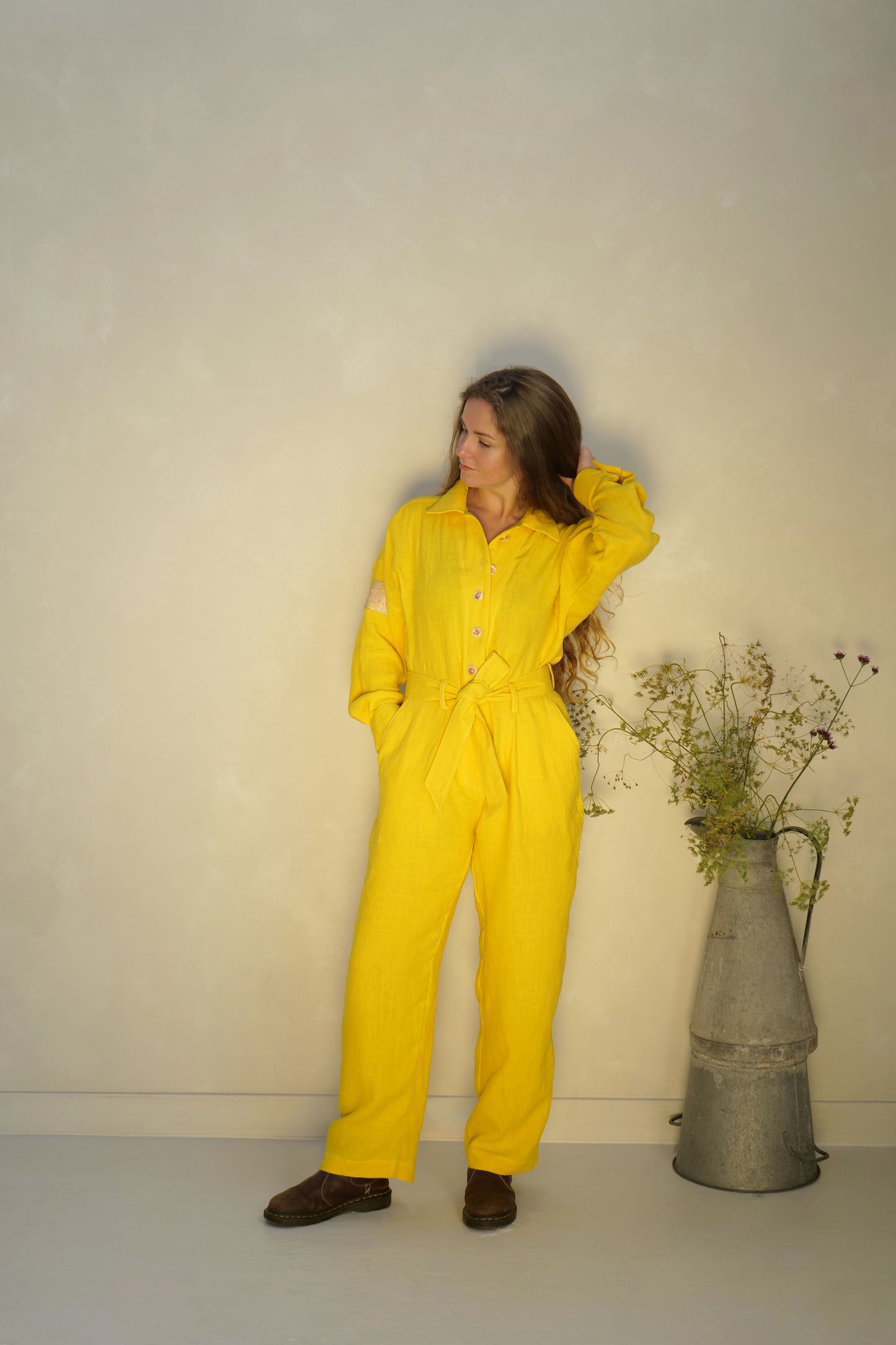 Person wearing a yellow linen jumpsuit standing next to a vase with flowers against a lime washed studio wall