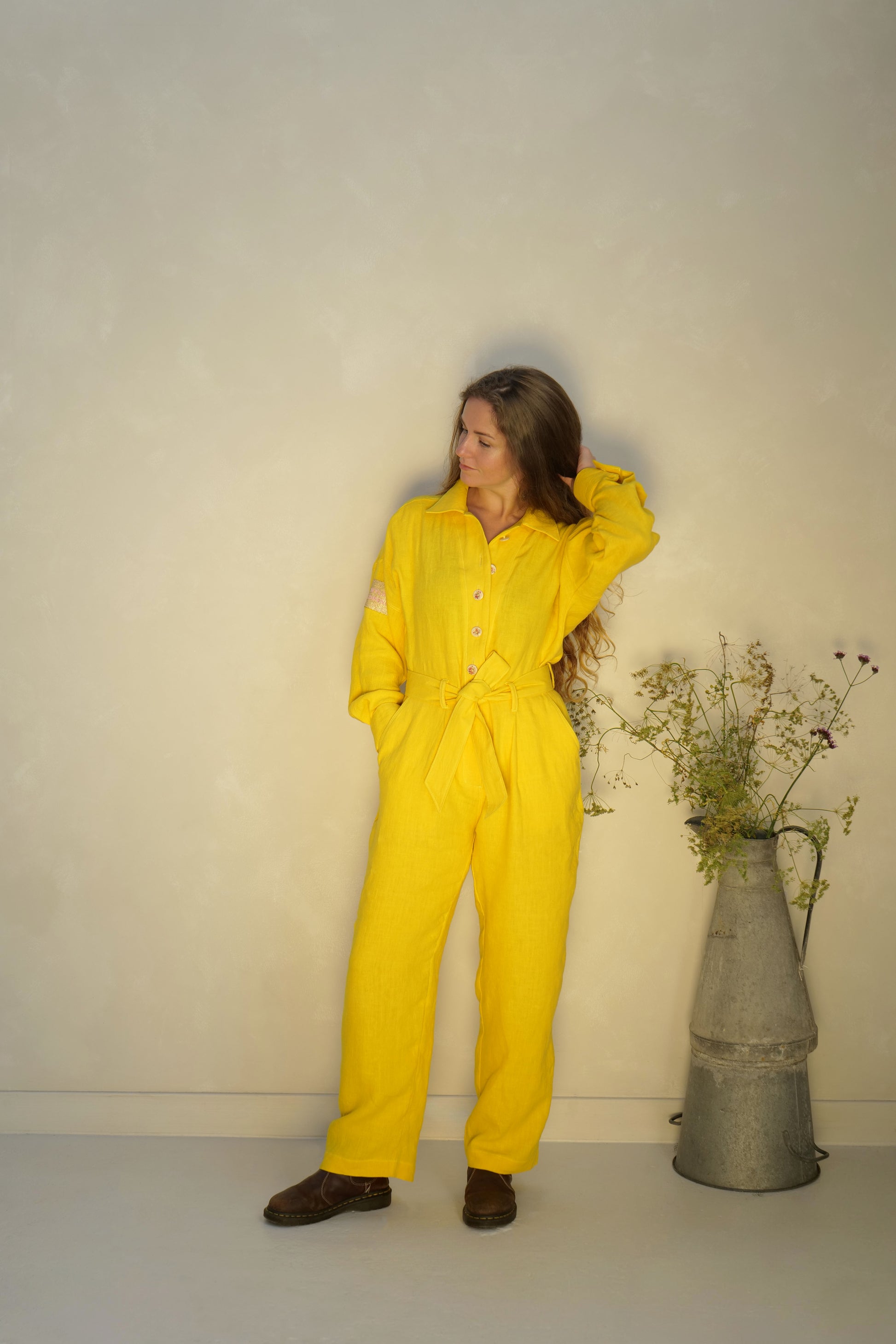 Person wearing a yellow linen jumpsuit standing next to a vase with flowers against a lime washed studio wall