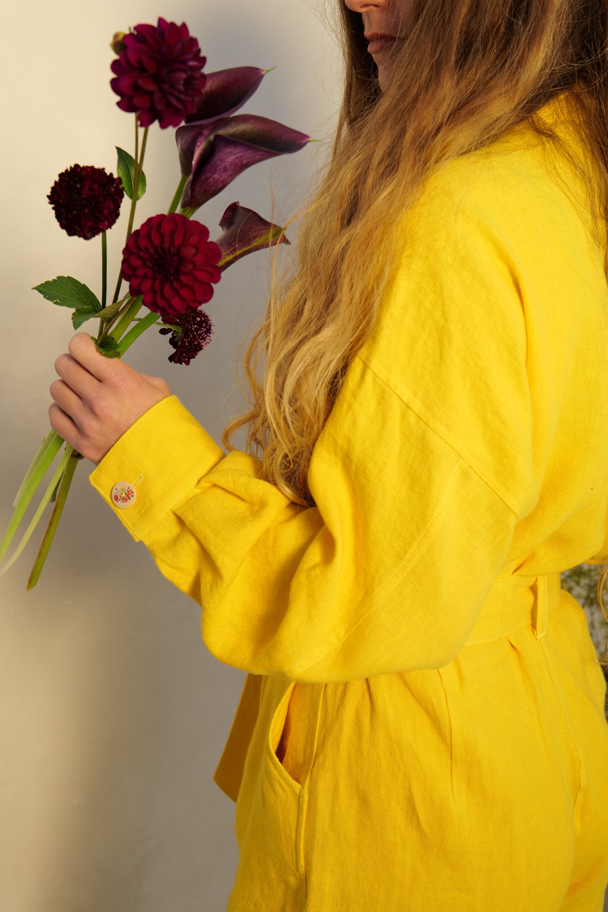 Person wearing yellow linen overalls holding dark red dahlias and calla lilies against a neutral background