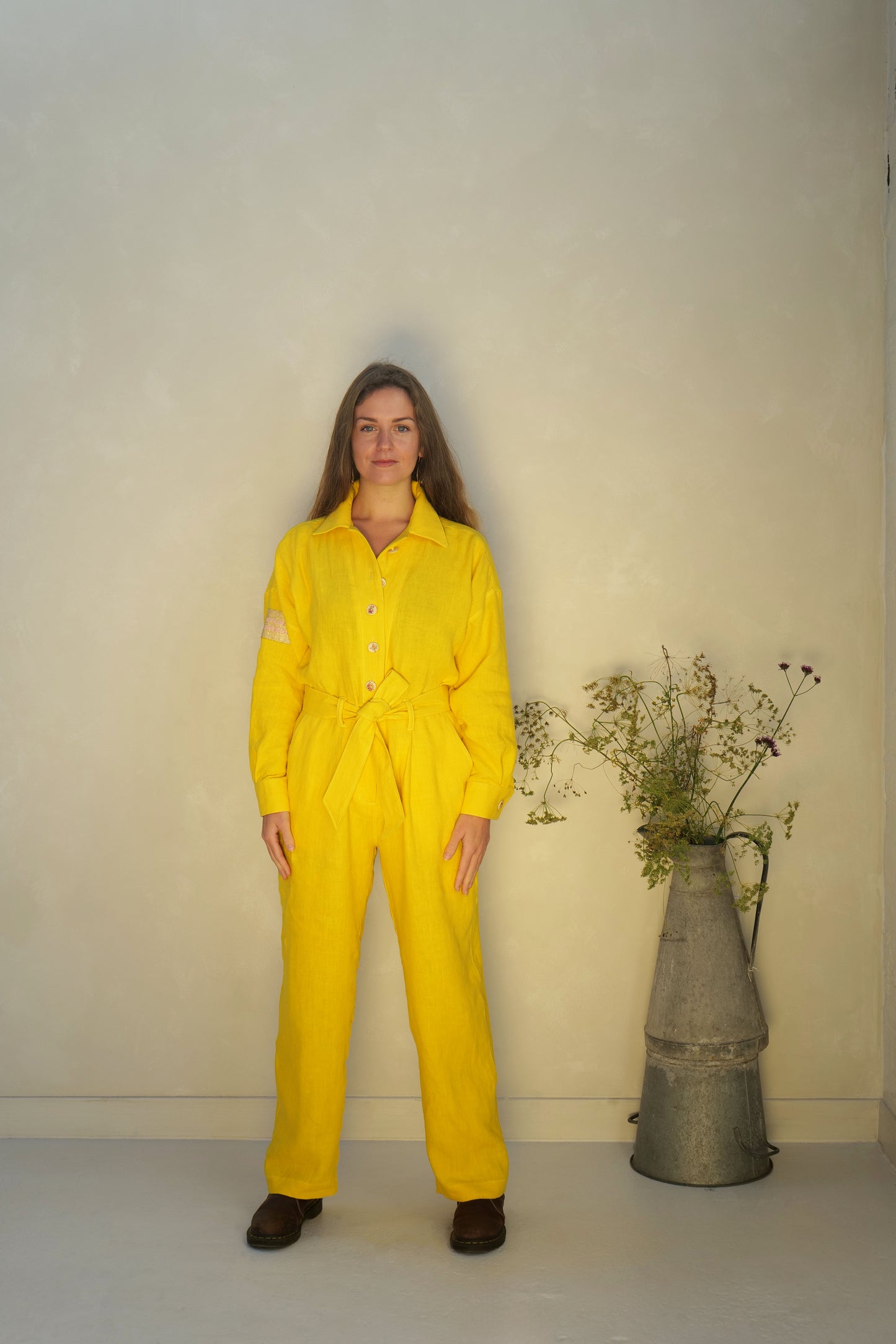 Person wearing a yellow linen jumpsuit standing next to a metal vase with plants against a plain background