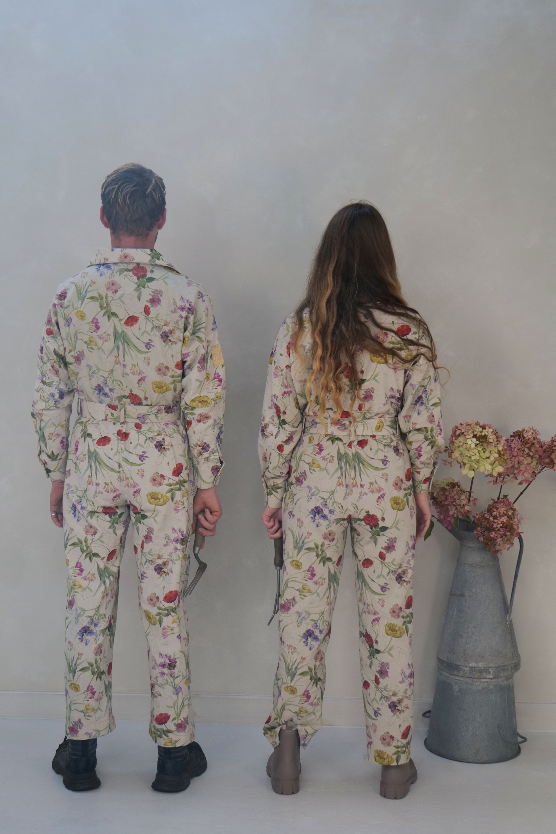 Two people wearing floral jumpsuits standing in front of a plain wall with a vase of flowers.