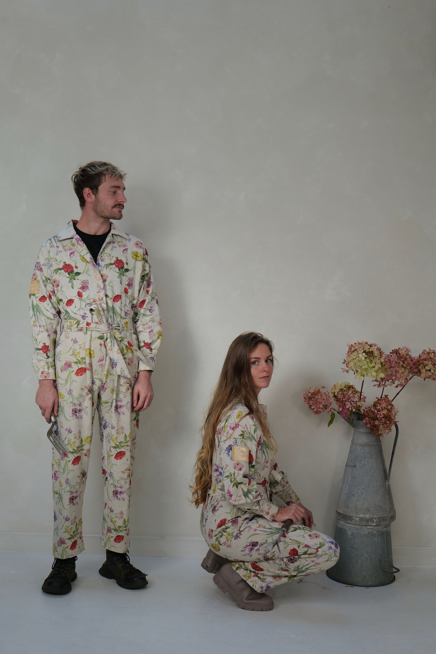 Two people wearing floral jumpsuits standing against a lime washed background with a vintage zinc jug of flowers.