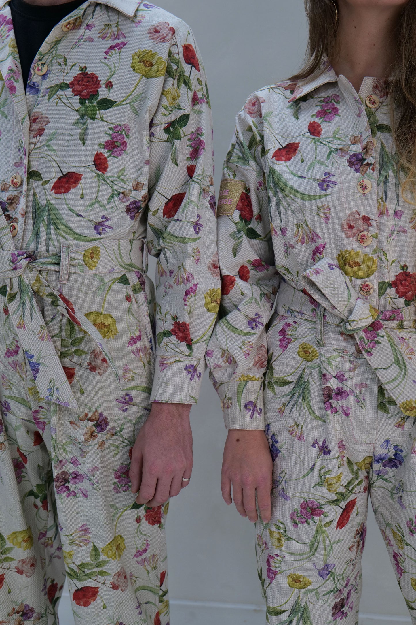 Two people wearing floral overalls against a lime washed background background