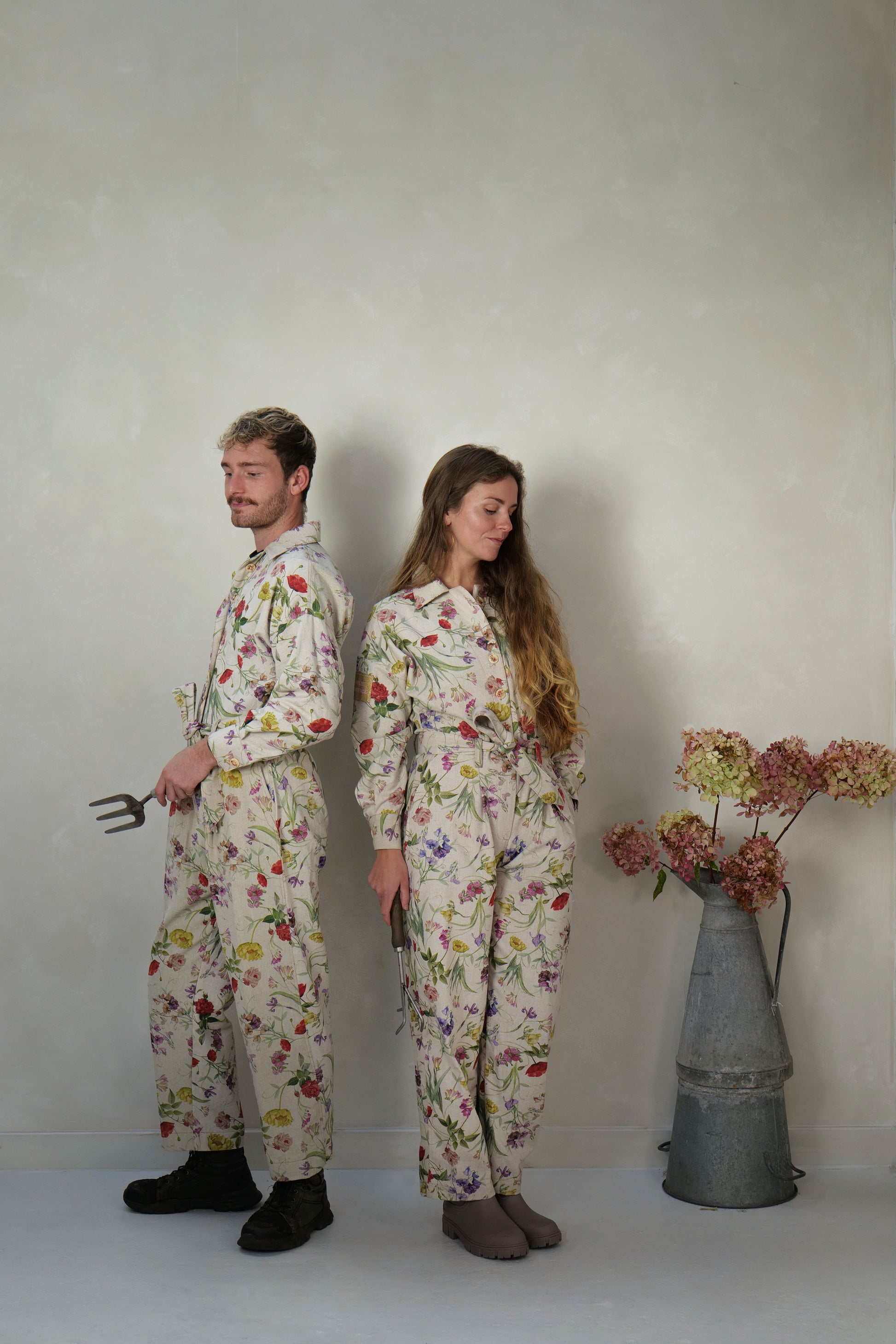 Two people wearing matching floral jumpsuits standing against a plain background with a vase of flowers.