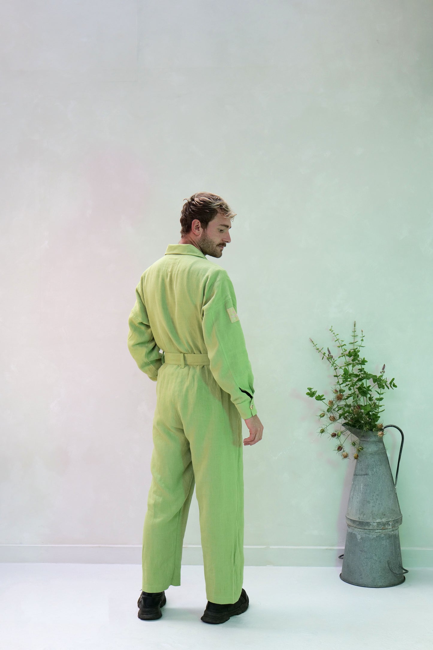 Man in a light green linen jumpsuit standing next to a vase with plants against a white background