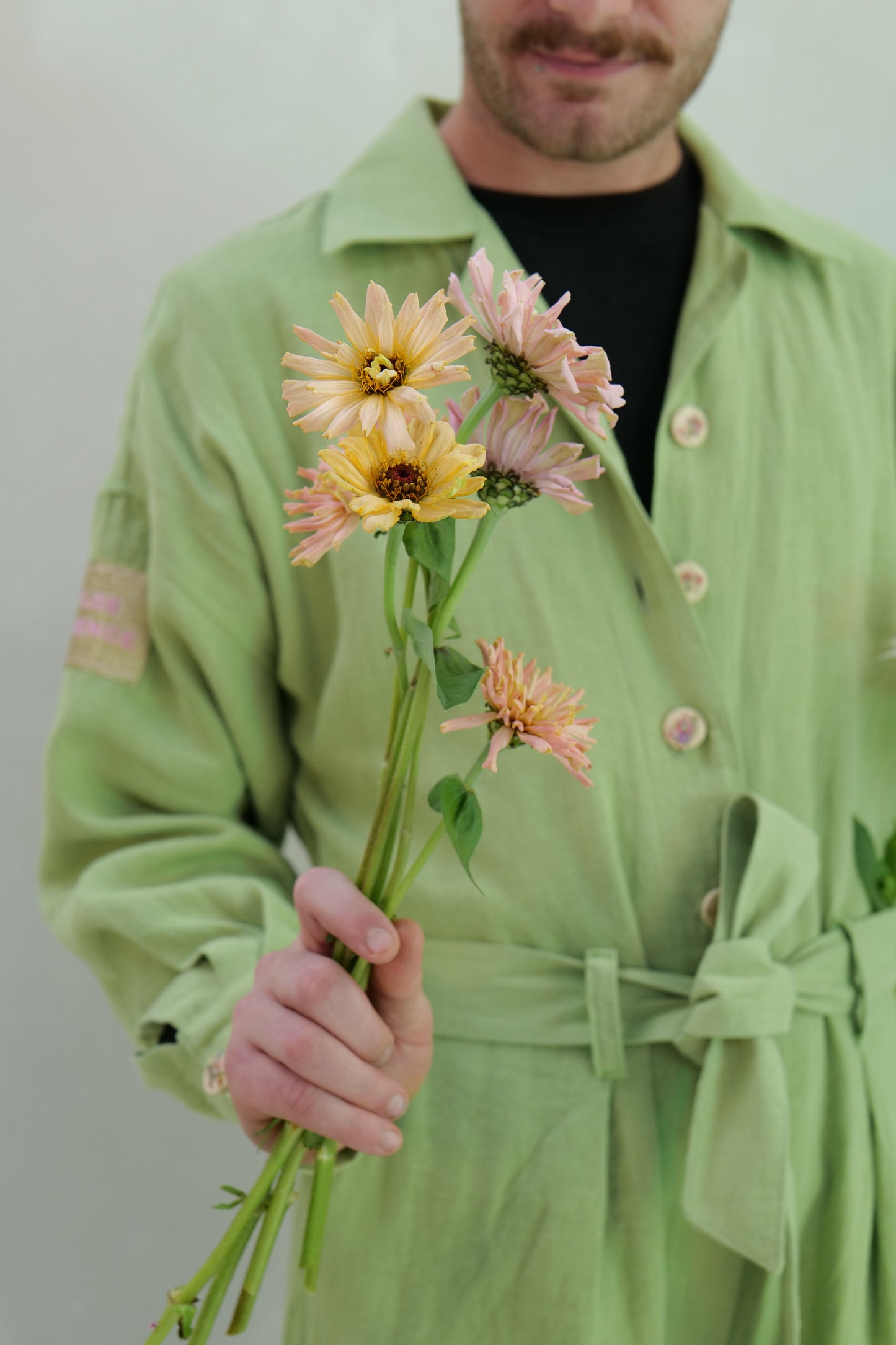 Person wearing light green linen overalls holding a selection of floret zinnias against a plain background