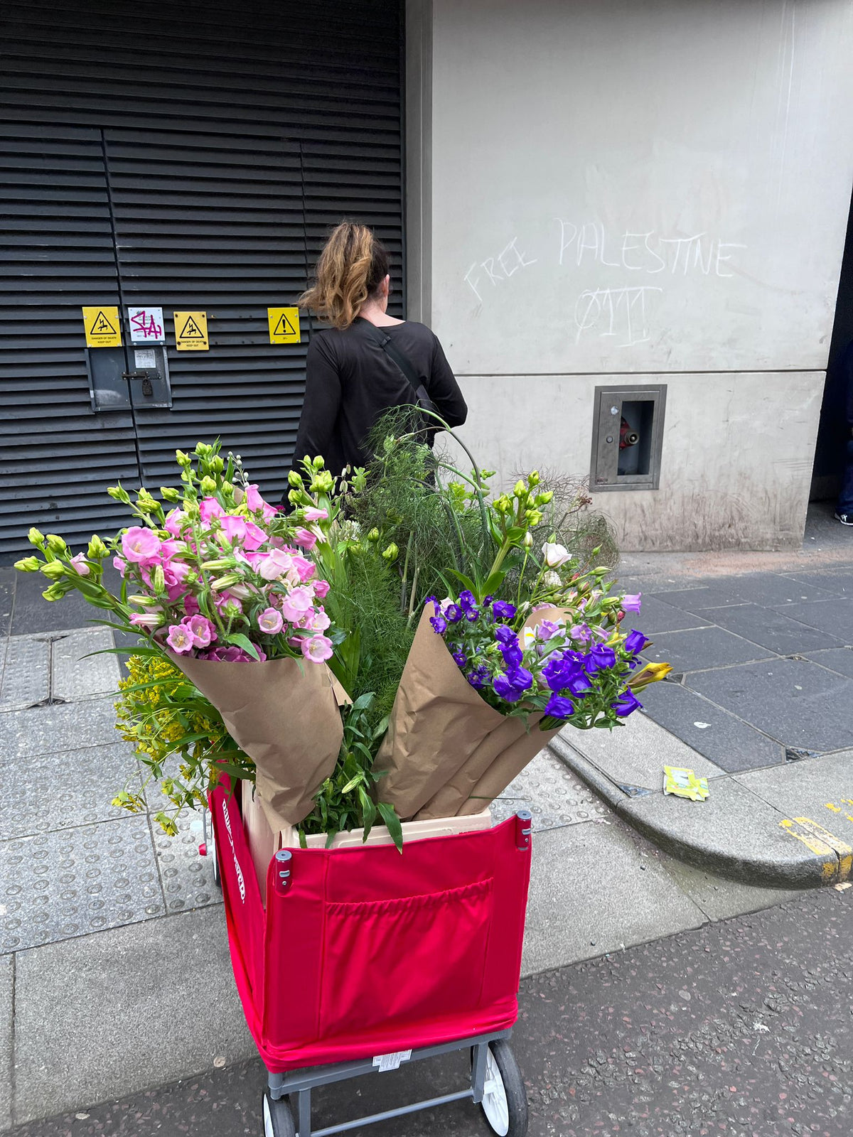 A red wheeled utility cart filled with assorted fresh flowers wrapped in brown paper, parked on a city sidewalk in front of a gray building wall with graffiti.