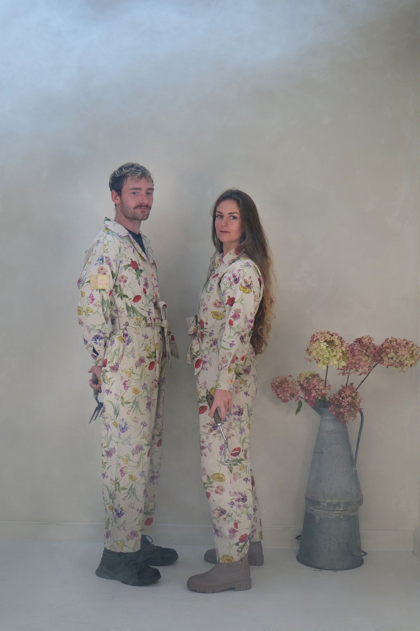 Two people wearing floral overalls standing in a studio with a vase of flowers.