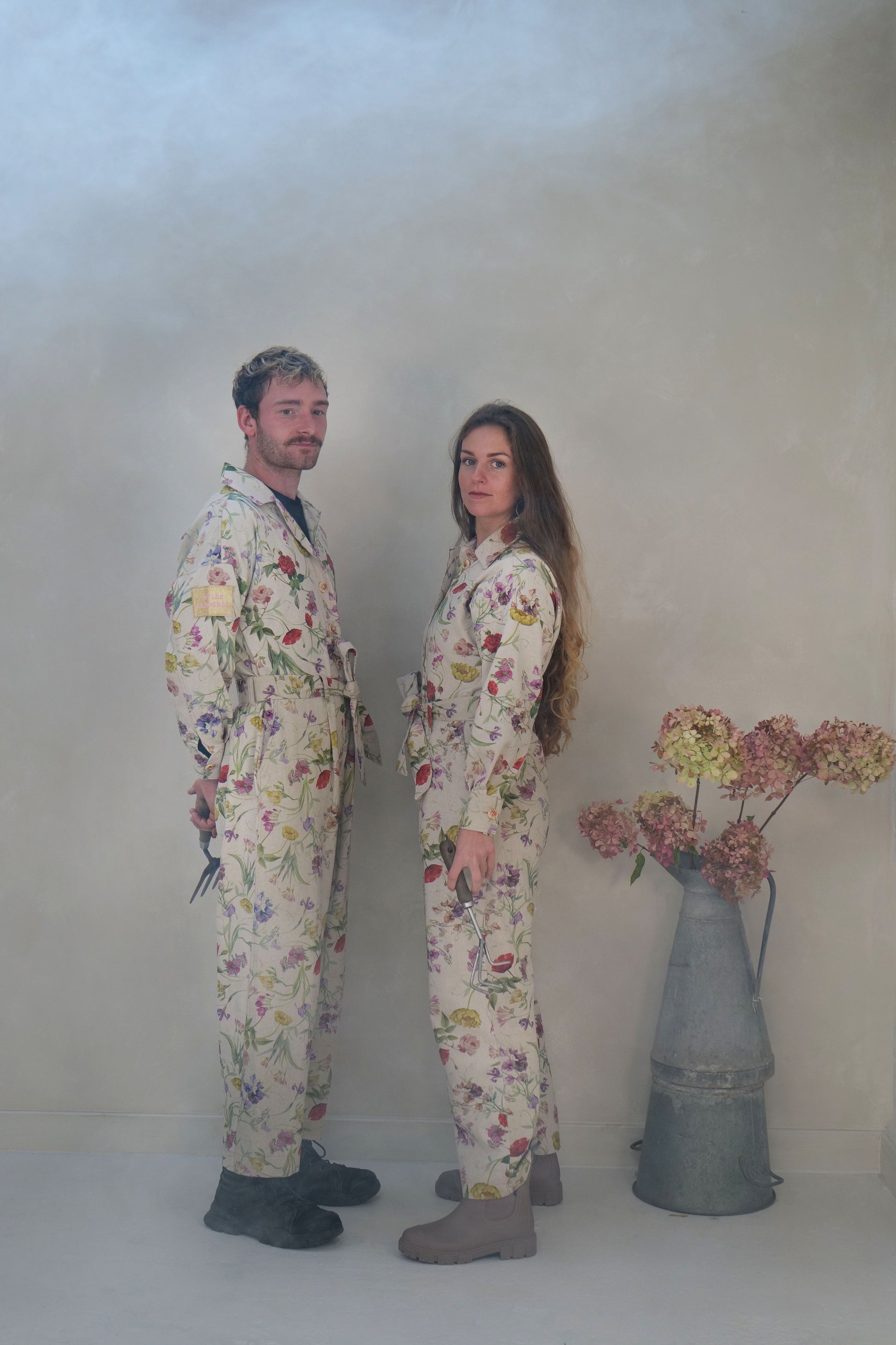 Two people wearing floral overalls standing in a studio with a vase of flowers.