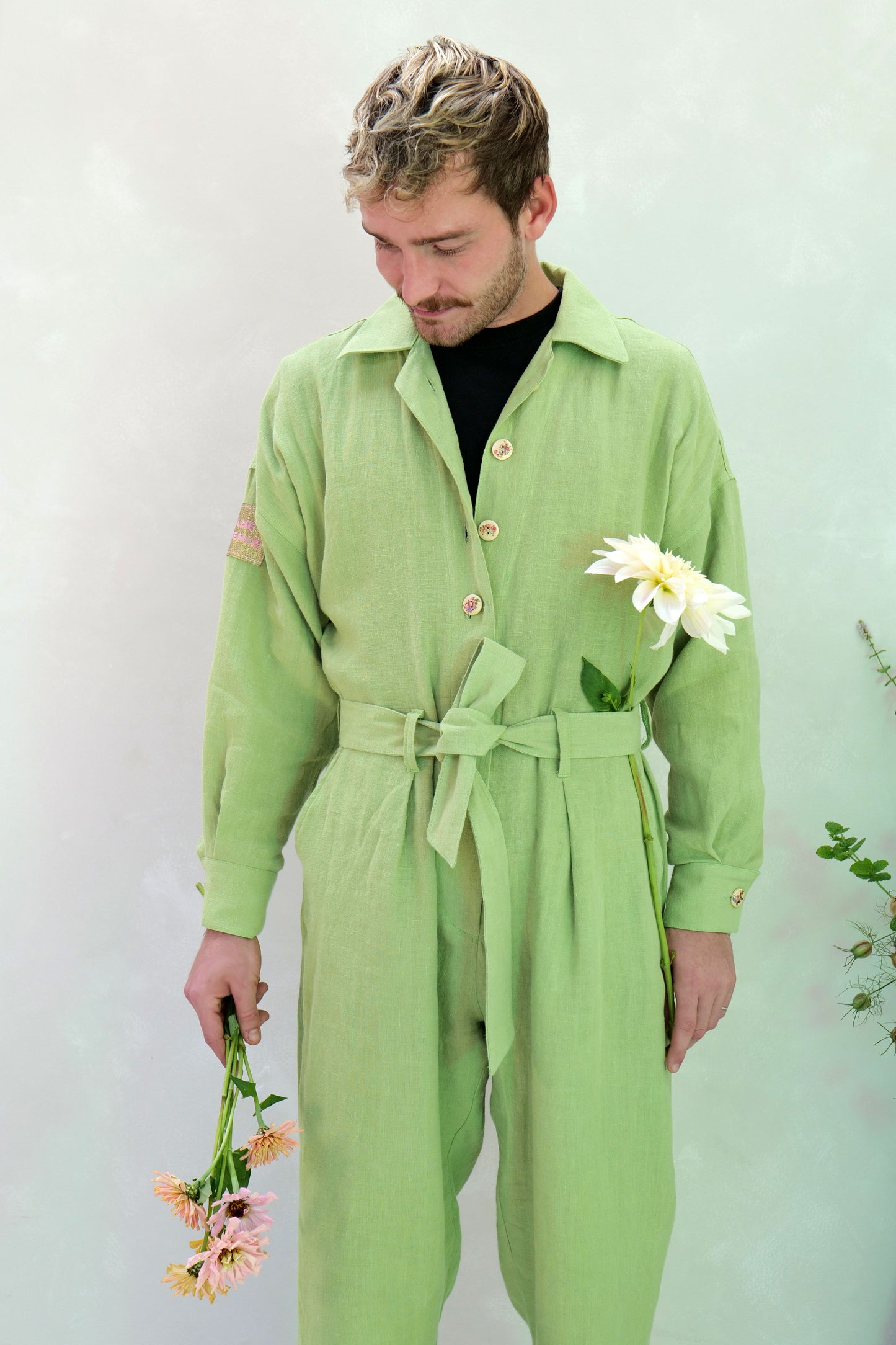 Man wearing a green linen jumpsuit holding flowers against a light background